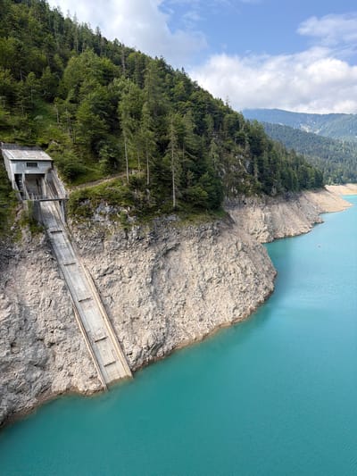 This is a high-angle, full shot of a concrete spillway channel descending from a building built into a steep, forested mountainside towards a vibrant turquoise body of water. The building at the top of the spillway is rectangular with a grey, angled roof and a single window visible. The spillway itself is a long, sloped, concrete structure with horizontal support beams visible along its length. The mountainside is covered in dense, dark green coniferous trees. The water is calm with slight ripples on its surface. The shoreline is rocky and uneven, with evidence of a dry bed above the current water level. In the distance, more forested mountains are visible under a partly cloudy blue sky. There are no people or animals visible in the image. The scene depicts a man-made structure integrated into a natural mountain landscape, likely a dam or reservoir spillway.