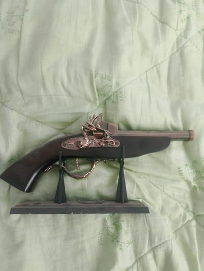 A vintage-style flintlock pistol replica is displayed on a dark rectangular stand, which is situated on a light green, quilted textile surface. The textile has a repeating pattern of bamboo shoots and the word "BAMBOO TEXTILE" printed in a light green color. The pistol has a dark brown wooden grip and a metallic barrel and mechanism, which appears to be brass or a similar alloy. The flintlock mechanism is highly decorative, featuring intricate metalwork. Two dark green, triangular supports hold the pistol in place on the stand. The overall lighting suggests an indoor setting with diffused natural light.