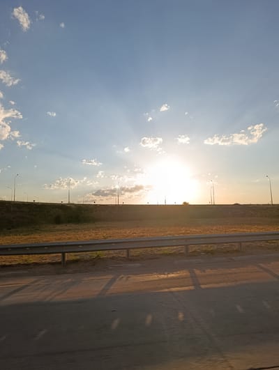 A wide-angle, eye-level shot captures a landscape scene on a clear day with a bright sun shining. The sky, a vibrant blue, is filled with scattered white clouds that are illuminated by the sun's rays, creating a dramatic sunburst effect. In the distance, a long, elevated highway runs horizontally across the frame, lined with streetlights at regular intervals. The highway is bordered by a grassy slope and a metal guardrail. The foreground of the image shows an asphalt road surface with long shadows cast upon it, suggesting the sun is low in the sky, possibly during sunrise or sunset. The overall impression is one of an open, possibly rural or semi-urban, setting bathed in natural light. The image is devoid of any human presence or activity.