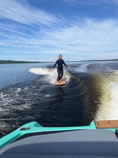 A woman in a black wetsuit surfboards on a wake behind a boat on a calm body of water. The boat is in the foreground, with a turquoise and gray interior visible. The woman is standing on a wooden surfboard, balancing as she rides the wave. She has blonde hair and is looking forward. The background shows a treeline and a blue sky with wispy white clouds. The location is Gromovo, Russia.