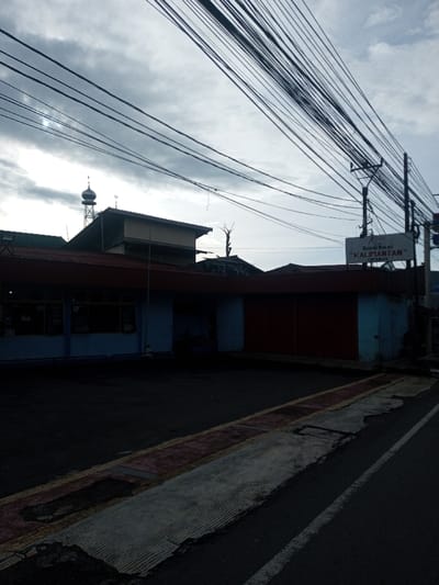 A street view shows a low, elongated building, painted blue and red, with closed shutters. Overhanging power lines crisscross the overcast sky. In the background, a mosque's dome is visible. A sign reads "Rumah Makan 'KALIMANTAN'", presumably a restaurant. The street's edge is partially visible, exhibiting cracks and disrepair. The scene is dominated by shadows, creating a somber atmosphere.