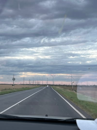 The image shows a road with a white line down the middle, bordered by a green field on the left and a line of wind turbines on the right. The sky is cloudy with patches of sunlight, suggesting it is either sunrise or sunset. The wind turbines are tall and modern, with their blades visible. There are also some power lines and poles along the side of the road. The scene appears to be rural and open, with no people or buildings immediately visible. The image is taken from inside a vehicle, as evidenced by the interior frame on the right side of the image. The overall mood is calm and serene, with a sense of vastness. The location is Zimovniki, Russia.