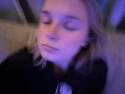 The image is a blurry, close-up shot of a person's face. The individual appears to be lying down, with the camera positioned at an angle. The lighting is predominantly purple and somewhat dim. The person's eyes are closed, and only the nose, mouth, and chin are partially visible. The background is indistinct, but seems to include fabric or clothing, as well as dark shadowy areas.