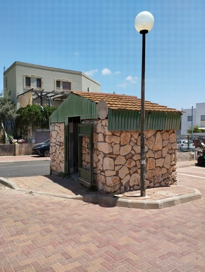 This is a photograph taken on a bright, sunny day in Zikhron Yaakov, Israel. The scene depicts a small, rustic building constructed with rough-hewn stone at its base and green corrugated metal siding under a brown tiled roof. The building has an open doorway with a green metal gate. To the right of the building, a tall, dark lamppost with a white, globe-shaped light fixture stands prominently against a clear blue sky with a few wispy white clouds.
In the background, a two-story building with light-colored walls and stone window frames is visible, partially covered by lush green foliage. A dark-colored car is parked in front of a garage door to the left of the stone building. The ground in the foreground is paved with reddish-brown brick pavers laid in a herringbone pattern. A grey curb separates the paved area from a street with asphalt.
A sign with Hebrew text is visible on the stone building, near the top left corner of the doorway. Another small, circular sign with Hebrew text is mounted on the green siding above the doorway. The overall impression is a sunny, warm day in a residential or public area in Israel, with a blend of traditional and modern architectural elements. There are no people visible in the image.