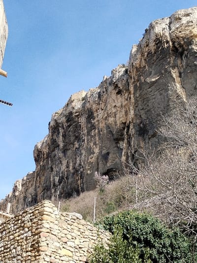A towering rock face dominates the scene under a clear blue sky. The cliff displays a layered, sedimentary structure with patches of dark staining. Bare trees and shrubs grow at the base of the cliff, interspersed with a few blooming trees. A stone wall sits in the foreground, built of rough-hewn rocks, with some green foliage draping over it. A partial view of a building is seen on the left.
