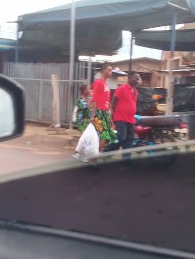 A medium shot shows three people walking in what appears to be an outdoor market area in Aketu-Oja, Nigeria. In the foreground, a motorcycle is parked. The area is paved with some debris scattered on the ground. A metal fence is visible behind the people. A white plastic bag is being carried by the person in the middle. Buildings are visible in the background, suggesting an urban environment. The lighting suggests it is daytime.