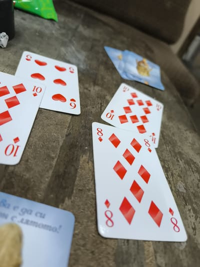 The image shows a close-up, oblique shot of several playing cards scattered on a wooden surface. The cards are primarily red, indicating they are likely from the diamond suit. Visible card values include '9', '10', and '8', with multiple diamond symbols depicted. There is also a card with a blue background and an image of a creature or person on it, partially visible. A partially obscured card with writing in a non-English alphabet is visible in the bottom left. The surface appears to be a rustic wooden table. The lighting suggests an indoor setting with a slight shadow cast. The image quality is slightly blurred, particularly on the background elements. There are no people visible in the frame. The setting does not provide any specific indicators of the city of Kalchevo, Bulgaria.