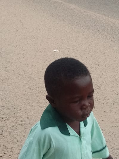 A young, dark-skinned child in a light green button-down shirt with a dark green collar stands on a light brown, textured surface. The child is looking down and slightly to the right. The background is an expanse of similar light brown, textured material. This image was taken in Biliri, Nigeria.