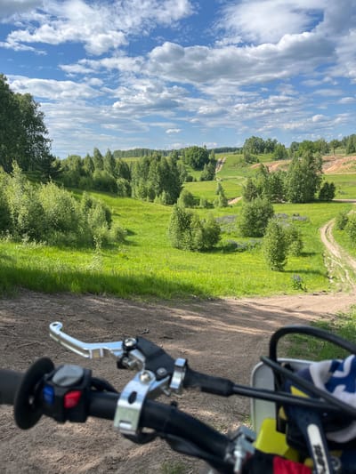 The image presents a first-person perspective from a stationary motorcycle or dirt bike positioned on a dirt track in Новочувашский, Russia. The handlebars of the vehicle occupy the lower portion of the frame, showing a silver clutch lever, a black handgrip, and a control module with red, blue, and grey buttons on the left. Partially visible on the right are a yellow and white section of the bike's front fairing, a black handgrip, and a blue and white patterned cloth draped over a cable.

The background reveals a vibrant, undulating landscape under a bright blue sky filled with numerous white cumulus clouds. Extensive green grasslands and numerous deciduous trees and bushes cover the hills and valleys. Multiple narrow, light-brown dirt paths or tracks wind through the grassy terrain, suggesting an off-road course or network of trails. Patches of purple flowers, likely lupines, are visible interspersed within the green grass, particularly in the midground to the right. To the right, an area of exposed, lighter-colored earth (possibly sand or clay) indicates a geological feature or excavation. No individuals are visible in the scene. The overall impression is a sunny, pleasant day in a natural, possibly recreational, outdoor environment.