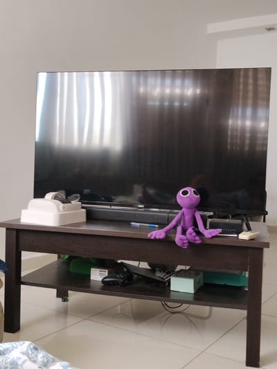 In what appears to be an indoor living room setting, likely in Be'er-Sheva, Israel, a large, dark television screen dominates the background, reflecting vertical light patterns from what may be blinds or curtains. Positioned on a dark wood coffee table in front of the television is a plush purple toy with large white circular eyes and three-fingered hands and feet. To the left of the toy, on the same table, is a white electronic device with a handle. The coffee table has a lower shelf where various items are visible, including what seems to be electronic equipment and boxes. The floor is tiled with a light-colored pattern. The overall scene suggests a domestic environment, possibly a living room area where entertainment devices are present.