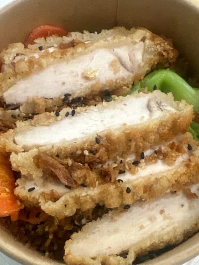 A close-up, indoor shot shows a serving of sliced, breaded chicken in a takeout container. The chicken is garnished with black sesame seeds. A piece of green lettuce and a few orange carrot slices are visible beneath the chicken. The container itself is a light brown color. The scene appears to be in France. The name of the city in the prompt is Tremblay-en-France.