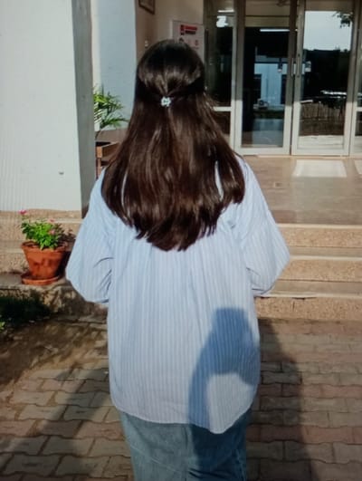 An image taken in Ortiugil, Uzbekistan, displays a solitary individual viewed from behind, positioned on brick paving, facing a building entrance. The person exhibits long, dark brown hair styled in a half-up configuration, secured by a small, light-colored claw clip. They are dressed in a light blue and white vertical striped long-sleeved shirt and a light blue denim skirt. A shadow is cast from the individual, visible on their shirt and the paving below. Three light-colored steps ascend to the building's entrance, which features two sets of double glass doors with dark frames. These doors reflect portions of the outdoor scene and provide a partial view of a dimly lit interior. To the left of the steps, a terracotta pot containing a green plant rests on a saucer on the ground, adjacent to a plain light-colored wall. A larger green plant is also visible further to the left, behind the potted plant. The scene is illuminated by natural light.