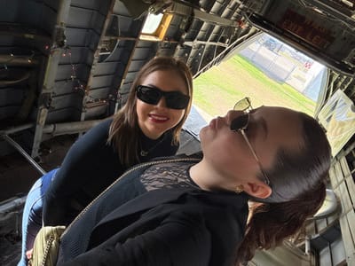 Two women pose inside what appears to be the interior of an old airplane. One wears sunglasses and a black top with lace detailing. The other has her hair pulled back and also wears sunglasses. The plane's interior is stripped down, revealing metal beams and sections. A window is visible in the background, offering a glimpse of a grassy area and what looks like an airport runway.