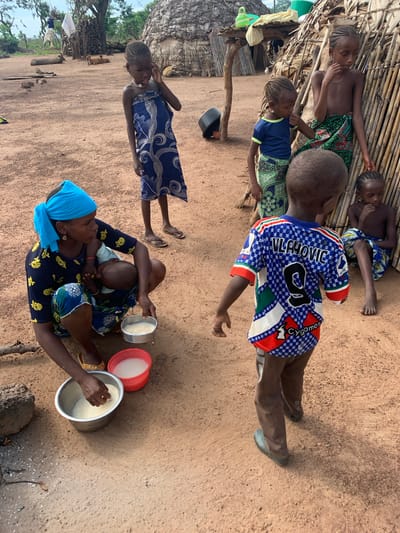 In the city of Nandu, Nigeria, a group of children and a woman are gathered in a dusty, outdoor area. A woman, wearing a blue headscarf and a dark blue dress with yellow floral patterns, is kneeling on the ground, breastfeeding a baby. She has a silver bowl of white grain in front of her, and her right hand is reaching into the bowl. Next to it, a red bowl contains a white liquid, likely water.
Around the woman, five children are visible. One young girl, wearing a blue and white patterned wrap, stands to her right, looking down and touching her ear. Another girl, wearing a blue t-shirt and a patterned green and blue skirt, stands to the left of a hut made of reeds and thatch, her hand resting on the shoulder of a boy in front of her. This boy is wearing a blue and white soccer jersey with "VLAHOVIC" and the number "9" on the back, along with "Cygames." He is facing away from the camera. To the right of the reed hut, another young child, a boy, is seated, looking down.
The environment consists of dry, reddish-brown dirt ground. In the background, a thatched hut with a woven reed wall is prominent, along with stacks of firewood and sparse vegetation. The scene appears to be a rural village setting.