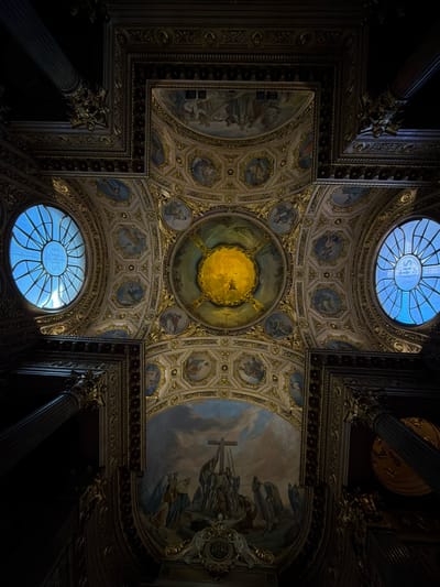 The image depicts the ornate ceiling of the Duomo di Santa Maria Assunta in Mozzo, Italy. The ceiling is richly decorated with frescoes and gilded architectural elements. Two large, circular stained-glass windows with intricate patterns are visible on either side, emitting a soft blue light. In the center of the composition, a large fresco depicts a religious scene, possibly the crucifixion, with figures gathered around a cross under a dramatic sky. The overall impression is one of grandeur and historical significance, indicative of a sacred and architecturally significant space.