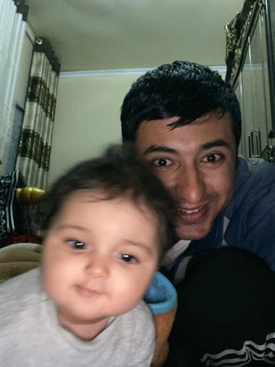 The image displays an adult male and an infant in an indoor setting, identified as Galaosiyo, Uzbekistan. The adult male, positioned behind and to the right of the infant, has dark hair, dark eyes, and a wide smile. He wears a blue top with a white collar and dark pants with white stripes on the side. The infant, located in the foreground and slightly out of focus, has dark hair and dark eyes, with a neutral expression and gaze directed slightly to the left. The infant wears a light-colored long-sleeved top, with blue and tan/brown fabrics visible around its chest and shoulder area. The background features light-colored walls. To the left, a window or opening is covered by vertical striped curtains (brown and beige/gold), with a gold-colored finial visible. To the right, an ornate dark object with gold accents, possibly part of a mirror frame or furniture, is partially visible.
