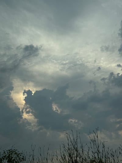 This is a vertically oriented, somewhat underexposed photograph of a dramatic sky. The scene is dominated by a dense, textured cloud cover, with areas of lighter, brighter clouds interspersed with darker, more ominous formations. A sliver of sunlight peeks through a gap in the darker clouds, casting a soft, warm glow on the sky. On the far right side of the frame, the thin, bare branches of a tree are silhouetted against the sky, suggesting a season of dormancy, possibly autumn or winter. The overall mood is one of impending change, perhaps a storm or the transition from day to night. The location is identified as Brashlen, Bulgaria. The time of day is ambiguous due to the lighting conditions, but the contrast between light and dark clouds could suggest either dawn or dusk. There is no visible text in the image.