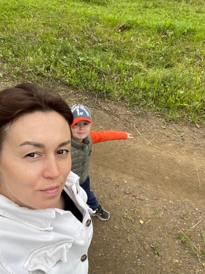 In Bormist, Russia, a woman and a young boy are captured in a selfie-style photograph. The woman, in the foreground and taking the picture, wears a white jacket. Behind her, the boy, wearing an orange long-sleeved shirt and a green vest, points his right arm forward. He has on a blue and red baseball cap with white lettering. They are outdoors in a dirt path area adjacent to a grassy field with sparse wildflowers. The background consists of the grassy terrain.