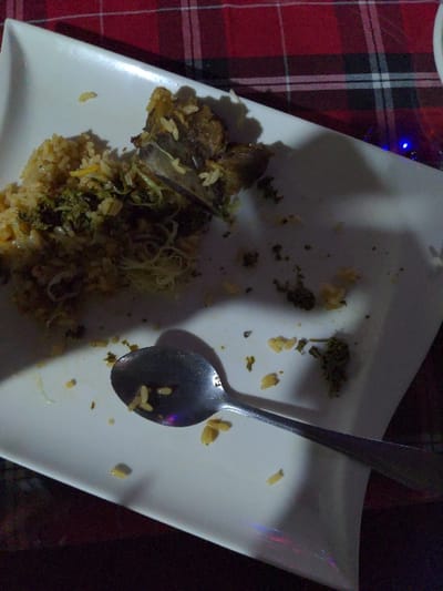 The image shows a close-up, overhead view of a white, rectangular plate with remnants of a meal, resting on a red and black plaid tablecloth. The plate is largely empty, indicating that the food has mostly been consumed. Scattered across the plate are grains of rice, some dark pieces of what appears to be meat or fish, and green, leafy fragments, possibly herbs or vegetables. A metal spoon lies diagonally across the plate, with a few grains of rice adhering to its bowl.
The lighting in the image is dim, casting soft shadows on the plate and creating reflections on the spoon, suggesting an indoor setting, likely in the evening or at night. A small blue light, possibly from a decorative string light, is visible in the upper left corner, partially obscured by the plate's edge. The overall scene conveys the aftermath of a meal. There is no visible text in the image. While the provided context states Bagamoyo, Tanzania, there are no specific visual cues within the image to confirm this location.
