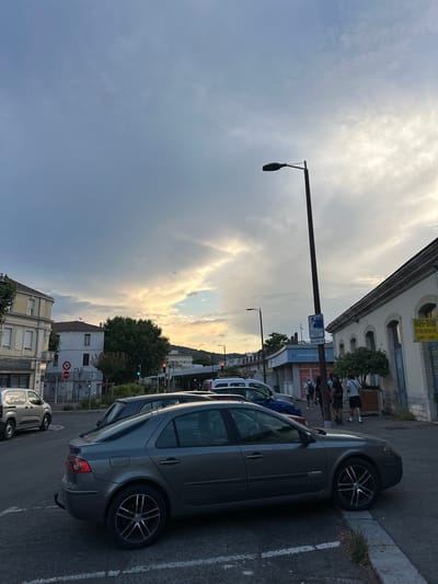 The image depicts a street scene in Alès, France, likely during late afternoon or early evening, given the quality of light. The sky is dramatic with a mix of dark, heavy clouds and areas of bright, possibly sunset light breaking through.
In the foreground, a dark grey sedan, possibly a Renault Laguna, is parked facing right, slightly angled towards the viewer. Its rear wheel is partially on a raised curb. To its left, the back of a dark blue car is visible, and further to the left, a silver van is parked.
The background shows buildings lining the street. On the left, a light-colored building with multiple windows and balconies is prominent. To its right, a white building with a tiled roof stands. Across the street, a long, light-colored building with arched windows and a blue awning is visible. Signs on this building include "ALES-TAXI" with a phone number, and "HAVAS VOYAGES."
Several people are walking near the building with the blue awning. They appear to be a mix of adults and possibly older children, dressed in casual clothing. Traffic lights are visible; one red light is illuminated on the left, and a green light is illuminated on the right. A street lamp with a distinctive curved head stands tall in the middle ground, near the building with the blue awning. A red "STOP" sign is mounted on a pole near the white building. The overall environment suggests a commercial or transport hub area within the city.