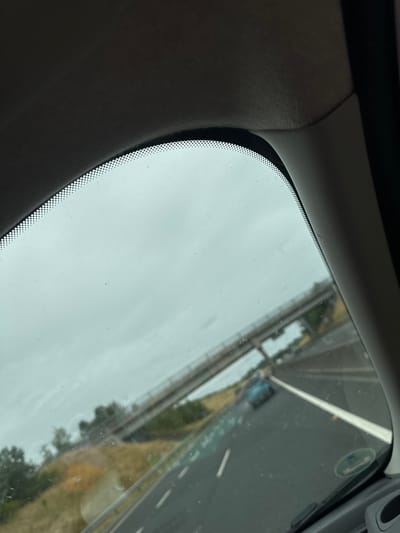 The image shows a view from inside a vehicle looking out through the windshield. The windshield has a dotted black border at the top. Outside, a cloudy sky is visible. A road with multiple lanes is seen, with white lane markings. There are cars on the road, including a light blue car in the distance. An elevated structure, possibly a bridge or overpass, crosses over the road in the background. The landscape next to the road appears to be grassy with some trees. There is no discernible evidence of the city of Moone, Ireland in the image.