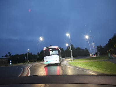The image captures a scene from within a vehicle, looking forward on a wet road at dusk. A bus, identified by the number 3458 on its rear, is traveling away from the viewer on a curved road. The bus has its brake lights illuminated, casting red reflections on the wet asphalt. Streetlights are visible along the roadside, illuminating the scene with their bright, white light.

In the background, a dense line of trees forms a dark silhouette against the twilight sky. A few other vehicles are visible further down the road, their headlights creating streaks of light. On the right side of the road, a bright green electric scooter is parked.

To the left of the road, a building with a dark facade and a wooden-looking roof is partially visible, with a blue and white striped bollard in front of it. Road markings, including red and white stripes, are clearly visible on the asphalt, indicating lane divisions or safety barriers. The sky is a deep blue, with some subtle cloud formations and what appear to be atmospheric distortions or light flares.

The overall impression is one of a typical evening commute or travel scene in a semi-urban or suburban environment, possibly on the outskirts of Tiskre, Estonia, given the context provided. The wet road suggests recent rainfall, adding a sense of dampness to the atmosphere.