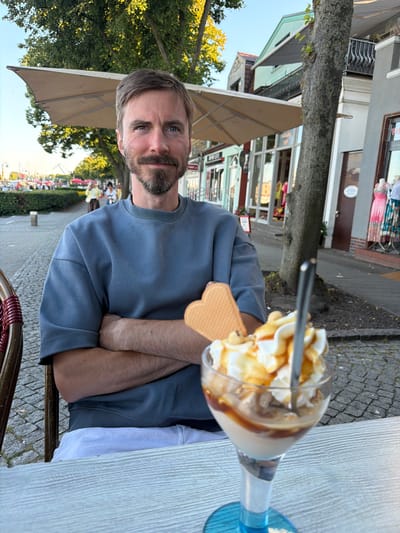 A man sits at a table with his arms crossed, looking at the camera. He has a beard and mustache and is wearing a blue sweatshirt. In front of him is a dessert in a glass, topped with whipped cream and caramel sauce, with a waffle on the side. The setting appears to be an outdoor cafe. The man is situated in front of a cobblestone sidewalk and a row of buildings. In the background, there are trees and people walking. This location is consistent with the promenade along the Warnow River in Rostock, Germany, a popular area with cafes and shops.