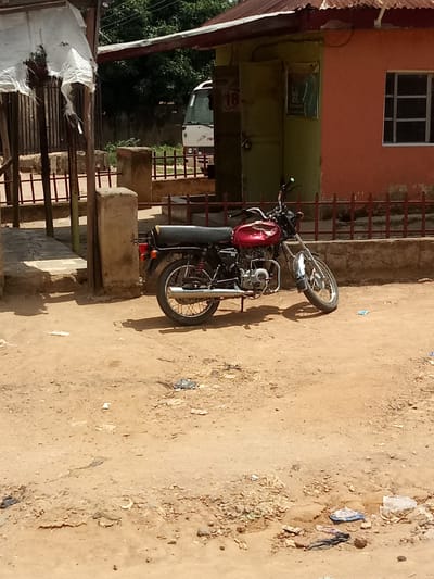 In Karshi, Nigeria, a red and silver motorcycle is parked on a dirt lot in front of a two-toned building. The building has an orange facade on the right and a green entrance area with a locked door on the left. A white bus is partially visible behind a metal fence. The motorcycle's black seat and red fuel tank are prominent. The ground is a mix of dry earth, scattered debris, and a few small plastic wrappers. A weathered concrete pillar stands to the left of the motorcycle. The background features trees and a corrugated metal roof.