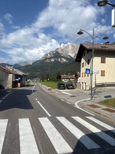 The image depicts a street scene in Mazzin - Mazin, Italy, on a sunny day with scattered clouds. A paved road with white lane markings runs diagonally from the foreground into the distance. A pedestrian crossing with white stripes is prominent in the lower portion of the frame. On the right side of the road, a light-colored building with brown shutters and a tiled roof stands. A street lamp with two lights is positioned in front of this building, along with a blue pedestrian crossing sign and a street sign that reads "Madonna de Lourdes". Two dark-colored SUVs are parked on the road in front of the building. Further down the road, more buildings and trees are visible, with a mountainous landscape covered in dark green trees dominating the background. On the left side of the road, a stone and wood building is partially visible, with a wooden fence and a blue pedestrian crossing sign. The overall environment suggests a quiet, picturesque village setting nestled in the mountains.