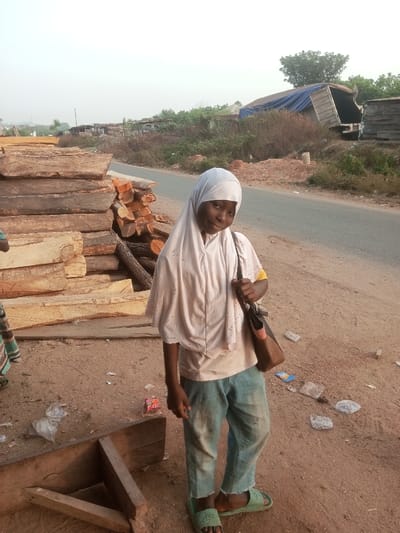 A young girl with brown skin, wearing a white hijab and casual clothes, stands on a dusty patch of ground. She is holding a brown bag. Behind her, a stack of logs is visible, and a road runs alongside the area. Trash litters the ground, including plastic bags and wrappers. In the background, a dilapidated structure with a blue tarp can be seen, adding to the overall impoverished setting.
