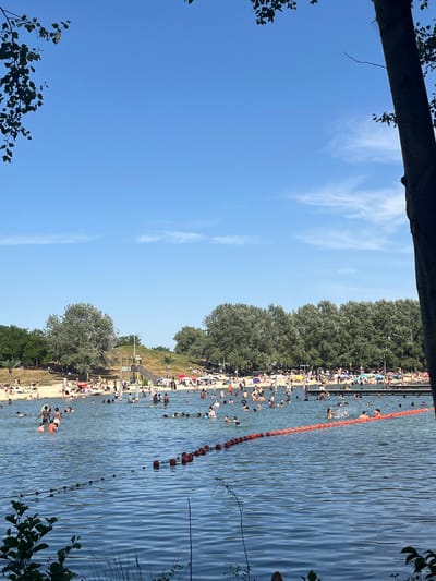 The image depicts a crowded outdoor swimming area at the municipal lake in Pomponne, France, on a clear, sunny day. The water is filled with numerous people swimming, wading, and playing. A red and orange buoy line separates a section of the lake. In the background, a sandy beach area is densely populated with sunbathers, many relaxing under umbrellas. Trees and foliage line the back of the beach, and a wooden structure with a ramp is visible. A large tree trunk is prominently featured on the right side of the frame, partially obscuring the view. The sky is a vibrant blue with a few wispy white clouds.