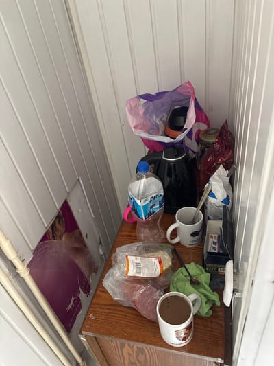 The image shows a close-up, high-angle view of a small wooden shelf against a white paneled wall. On the shelf are various items, including a black electric kettle, a bottle of "Aqua" brand water, a white mug with a logo and text that appears to be a phone number and location in Minsk, a bag of coffee, and other food items in plastic packaging. There is also a green cloth with a spoon on it and another white mug filled with a dark liquid, possibly coffee, with a Porsche logo on it. To the left of the shelf, partially visible, is a large poster with a woman's image and some text. The overall impression is that of a cluttered domestic or break area. The image does not contain any information that would indicate its location in the city of Vileyka, Belarus.
