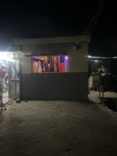 A small store at night with a brightly lit window displays merchandise. The building is a modest structure with a gray exterior. A wooden framework is visible below the window, and wires hang above. The surrounding area is dark and appears to be an unpaved alleyway or street. The overall scene depicts a simple, potentially rural commercial setting.