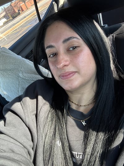 The image shows a young woman captured in what appears to be a selfie, seated inside a vehicle on a bright, sunny day. Her head is tilted, and she is looking directly at the camera with a soft, subtle smile.
She has long, straight, dark hair, likely black, parted on the side and flowing over her shoulders and chest. Her eyebrows are dark and well-defined, and she has brown eyes. Her complexion is fair with numerous freckles visible across her nose and cheeks. A small silver ring is piercing her left nostril. Her lips are slightly parted, revealing a natural pink hue.
Around her neck, she wears two golden necklaces: one is a rope-style chain, and the other is a finer chain with a small, sparkling pendant shaped like the letter "Y". She is dressed in a dark-colored (likely black or dark grey) crewneck sweatshirt. On the right side of her chest, partially obscured by her hair and the angle, there is white or light-colored text printed on her sweatshirt. Visible portions suggest words like "ART INSTITUT" or similar, with "AR" and "TUT" clearly discernible.
The setting appears to be the interior of a car. A dark headrest is visible behind her head. Through the car window to her left, a view of a road with yellow lines and distant buildings or trees can be seen, indicating an outdoor setting during the day. Strong sunlight is present, casting shadows on her hair and clothing. Next to her, on what seems to be the car seat, there is a fluffy, cream-colored object, possibly a blanket or a seat cover.