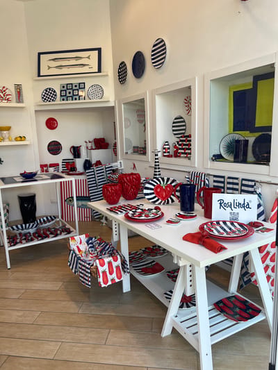 The image depicts the interior of a retail establishment, likely a shop specializing in tableware and home decor, located in the city of Sorrento, Italy. The store is brightly lit, with white walls and a wooden floor. Several display shelves and tables are arranged throughout the space, showcasing a variety of items.
The merchandise is predominantly red, white, and blue, with many pieces featuring nautical or food-related motifs, such as fish, tomatoes, and stripes. Notable items include ceramic plates, bowls, pitchers, vases, and decorative objects. A prominent display on the right features a sign with the text "Rosa Linda" and a QR code.
On the left side of the image, a wall display includes framed artwork of two fish, along with several decorative plates. A white shelving unit below holds more plates and a tall black vase.
In the center and right of the image, two white tables are set up with more tableware. The front table has various plates, napkins, and pitchers. A basket filled with folded cloths is placed on the floor in front of one of the tables.
The overall impression is one of a well-organized and visually appealing retail environment, emphasizing a specific aesthetic. There are no people visible in the image.