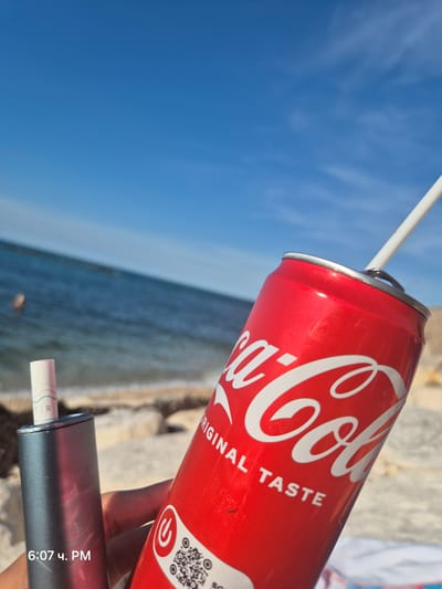 The image shows a close-up of a hand holding a red Coca-Cola can with a white straw and a metallic cylindrical object, possibly an e-cigarette, in front of a blurred background of the sea and beach under a clear blue sky. The time stamp "6:07 ч. PM" is visible in the bottom left corner. The scene appears to be a casual outdoor setting, likely a beach, on a sunny day. The location is the city of Bisceglie, Italy, as indicated by the user prompt.