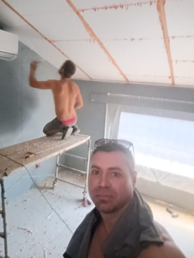 This media file depicts a domestic scene in Ladyzhyn, Ukraine, likely a renovation or construction project. In the foreground, a man is taking a selfie, wearing sunglasses on his head and a grey towel draped around his neck. He appears to be mid-task, with a light smattering of dust or debris on his shoulders. Behind him, a second man is seen working on the wall of what appears to be a room. He is shirtless, wearing grey pants and red underwear, and is kneeling on a scaffolding platform as he paints the wall. The room itself shows signs of ongoing work, with scattered debris on the floor and plastic sheeting covering a window, suggesting it's a work in progress. The ceiling has exposed wooden beams with orange sealant or paint applied. The overall impression is of a casual, potentially hot day, given the attire of the individuals and the bright light filtering through the window. The presence of an air conditioning unit on the wall suggests a controlled indoor environment despite the outdoor conditions.