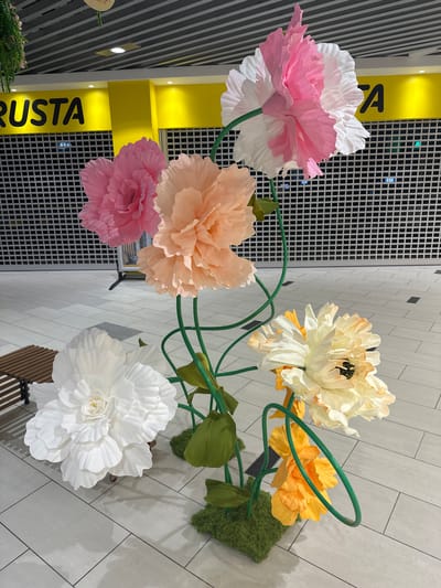 A top-down shot shows a display of large, artificial paper flowers with a modern, retail-like setting. The flowers are arranged on green wire stems that curve and loop, with some green moss accents. The flower colors include white, pink, pale orange, cream, and yellow. The background consists of a tiled floor in varying shades of grey and white, with a black tiled wall or partition that has a grid pattern. To the left, a bright yellow vertical sign with black text is partially visible. The visible text on the sign reads "STA" and part of another word. The lighting appears to be from overhead, consistent with an indoor retail environment, and there are no visible people. The overall impression is a decorative display in a public space.