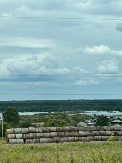 The image depicts a rural landscape with a cloudy sky. In the foreground, a large stack of hay bales is arranged in three rows, with each row containing approximately ten bales. The bales are cylindrical and appear weathered. Behind the hay bales, a body of water is visible, bordered by dense green vegetation and trees. A few structures, possibly houses or sheds, can be seen nestled amongst the trees near the water. The background consists of a dark, continuous line of trees, and beyond that, a pale blue sky filled with scattered clouds. The overall scene suggests a countryside setting, likely during the daytime given the lighting. There are no people or animals visible in the image. The location is identified as the city of Ключи, Russia.