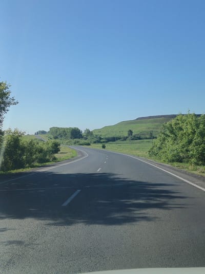 The image depicts a paved road curving through a grassy, hilly landscape under a clear blue sky. On the left side of the road, there are dense green bushes and trees. The road itself has a white dividing line. In the distance, there are more trees and the contour of a hill. The scene is likely captured during daylight hours, with shadows of trees cast on the road's surface. There are no people or vehicles visible. The location is identified as the city of Belovo, Russia.