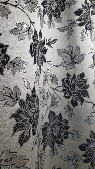 The image shows a close-up of a fabric with a floral pattern. The background color is a light silver or gray with a subtle sheen. The floral design consists of stylized flowers and leaves in shades of gray and black. The fabric appears to be textured and has a slight shimmer in some areas, suggesting metallic threads or a similar effect. The overall impression is of an elegant, possibly upholstery or drapery fabric. There is no evidence to indicate that the image was taken in Sannikovo, Russia.