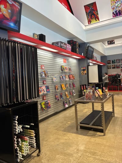 The image depicts the interior of a comic book store. A wall-mounted display of comic books is visible, organized on horizontal shelves against a grey, slatted background. Several comic books are displayed in clear plastic sleeves. To the left, a tall, black rack is filled with what appear to be rolled-up posters or comic book precursors, each individually wrapped and tagged. Above the comic book shelves, a red shelf displays various comic book boxes, including one labeled "SPIDER-MAN." A television screen is mounted on the wall, showing an image of Iron Man. The ceiling features white panels and some red accents. A metal display table with black shelves is positioned in the foreground, holding a few comic books and other merchandise. A young person wearing a grey hoodie and a black baseball cap is standing near the display table, facing away from the camera. In the background, more comic book artwork is displayed on the walls, and a rack of clothing is visible. The floor is a polished concrete surface. The scene is brightly lit.