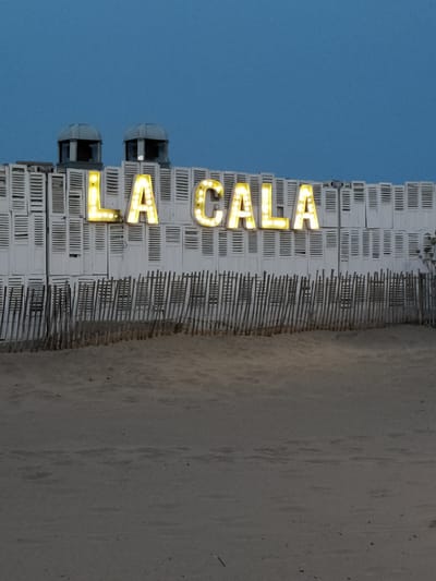 A beachside facade displays a prominent, illuminated sign spelling "LA CALA" in large, blocky, yellow letters with individual bulbs. The structure behind the sign is composed of stacked white shutters. Two domed structures with visible openings are situated atop the shuttered wall. In front of the building, a low fence made of thin wooden stakes separates the sandy beach from the structure. The scene is captured against a twilight sky of a deep blue hue.