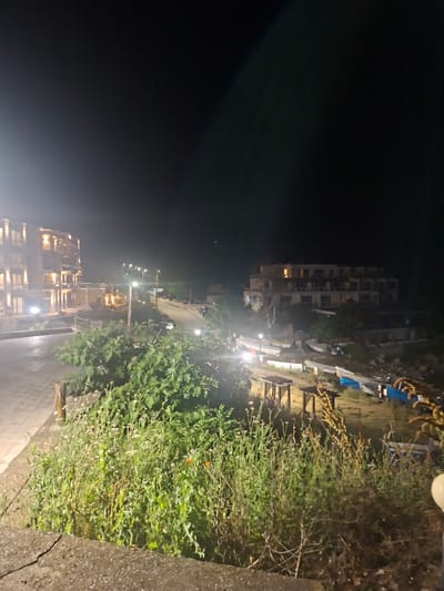 The image depicts a nighttime scene in Tyulenovo, Bulgaria, featuring a coastal resort area. In the foreground, dense green foliage with scattered small flowers grows along a cracked concrete embankment. Beyond this, a paved walkway leads towards two buildings. The building on the left is a multi-story structure with many illuminated windows, suggesting it is a hotel or residential complex. A lamppost with a bright light stands near the walkway.
Further into the scene, a road curves upwards, with a white van and at least one dark car visible. Several other buildings are spread across the landscape. To the right, a hill slopes down towards the coast. On this slope, several boats are resting on the ground, some covered with tarps. Two small, open-sided wooden shelters with slatted roofs are also present in this area. The background is dominated by the dark night sky, with a few distant lights suggesting the presence of more structures. The overall impression is one of a quiet, somewhat undeveloped resort town at night.