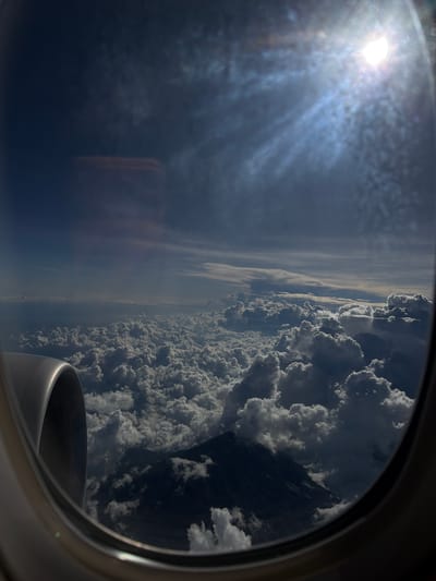 The image shows a view from an airplane window. Outside the window, there is a bright sun, scattered clouds, and a layer of haze. Below the haze, there's a dense cloud formation. Part of the airplane's engine is visible in the lower right corner of the frame. The location is not discernible from the image.