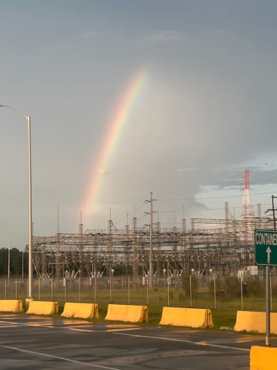 A rainbow arches across a cloudy sky above a large industrial electrical substation in Port Wentworth, Georgia, United States. The substation features numerous metal structures, wires, and poles. In the foreground, a line of yellow concrete barriers sits on the side of a wet asphalt road. A green sign with white text reading "CONTAINER" and an upward-pointing arrow is visible on the right side of the image. A tall white lamppost stands to the left, and a red and white tower is visible in the distance behind the substation. The sky is predominantly grey with some lighter areas where the rainbow is most prominent.