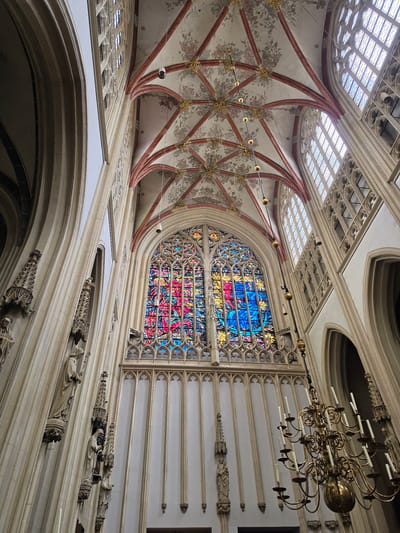 This image depicts the interior of St. John's Cathedral in 's-Hertogenbosch, Netherlands. The focal point is a large, ornate stained-glass window, showcasing a vibrant array of blues, reds, and yellows, likely depicting a religious scene. Above the window, the vaulted ceiling is adorned with intricate floral patterns in shades of pink and gray, bordered by red ribs and highlighted with gold accents. Several long, golden pendant lights hang from the ceiling.
Flanking the stained-glass window are tall, pale stone columns with Gothic architectural detailing. Niches carved into the columns contain several statues of robed figures, presumably saints, holding staffs and other religious symbols. To the right of the window, more stone architecture rises, featuring arched openings and more Gothic ornamentation.
In the lower right quadrant of the image, a large, multi-tiered brass chandelier hangs, equipped with numerous white candles, some of which appear to be lit. The overall impression is one of grandeur and historical significance, characteristic of a medieval European cathedral. The lighting is somewhat dim, allowing the stained glass to be a prominent source of color.