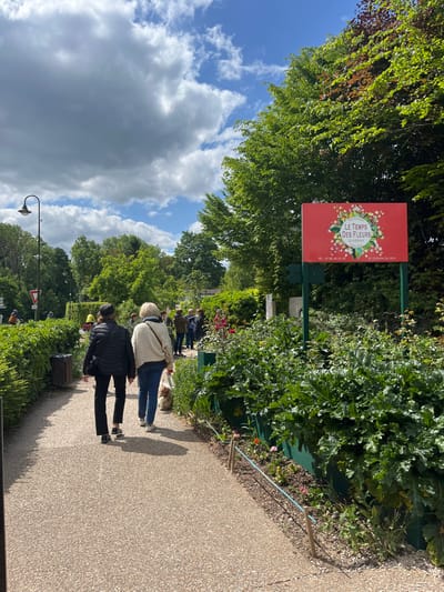 The image shows a gravel path bordered by green hedges leading toward a sign that reads "Le Temps des Fleurs Giverny" on a red background.  The sign is located at 49.075, 1.53. There are two women walking on the path. The woman in front is wearing a white sweater and black pants. She is walking in front of a woman wearing a black jacket and black pants. There are several other people standing to the right, in the distance. The sky is cloudy with visible blue patches.  There is a small, white dog walking in front of the woman in the white sweater. The ground is gravel and there is green foliage surrounding the path. The foliage to the right of the path is thicker than the foliage on the left.  There is a streetlamp to the left of the path.
