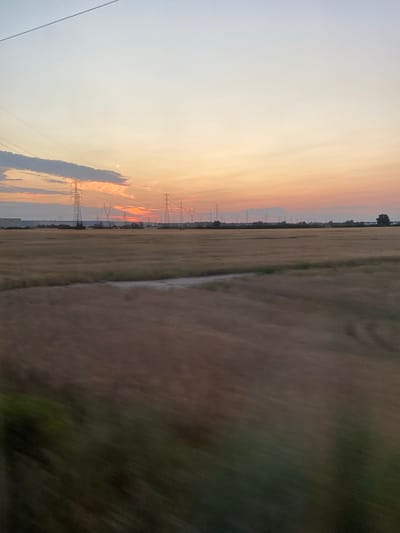 The image captures a sunset over a field with a line of tall electrical pylons in the distance. The sky transitions from a pale blue at the top to shades of orange and pink near the horizon, where the sun is setting. Wispy clouds are scattered across the sky, some catching the warm light. In the middle ground, the field appears to be covered in dry grass or crops, with a faint path or lighter colored strip running through it. Beyond the field, the electrical pylons stand tall, silhouetted against the colorful sky. Further in the background, a low-lying structure, possibly a warehouse or industrial building, is visible, along with a few scattered trees. The foreground shows a blurred area of green and brown, suggesting movement or a close-up of vegetation. The scene is peaceful and captures the natural beauty of a sunset in an open, rural or semi-rural landscape. It is likely taken from a moving vehicle due to the blur in the foreground.