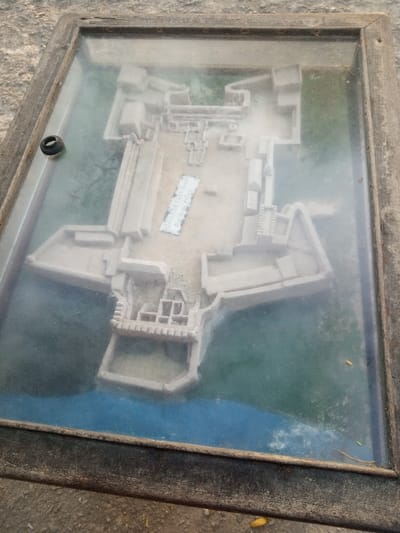 A weathered wooden-framed display case, covered by a dirty and reflective glass panel, is presented on a rough, light grey concrete surface. Inside the case is a beige-colored relief model depicting the layout of a historical fortification, identifiable as Fort Jesus in the Mji wa Kale ward of Mombasa, Kenya. The model features angular bastions, various rectangular structures, and internal courtyards. A significant portion of the model's perimeter, representing the surrounding environment, is colored dark blue for water and dark green for land/vegetation. A small, rectangular white label with faded, illegible black text is adhered within the central courtyard area of the model. A loose black rubber ring rests on the glass surface near the top-left corner. The wooden frame shows signs of aging and wear, secured by visible metal fasteners.
